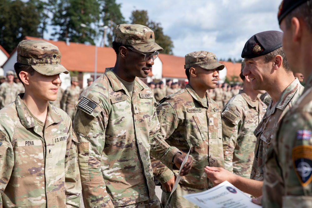 A military ceremony or award presentation is taking place outdoors, likely at a U.S. Army installation in Europe. A soldier identified as 'KNIGHT' is receiving or exchanging a document/certificate from an officer wearing a black beret with what appears to be a British or NATO insignia. Multiple U.S. Army soldiers in OCP (Operational Camouflage Pattern) uniforms are standing in formation in the background.
