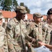 A military ceremony or award presentation is taking place outdoors, likely at a U.S. Army installation in Europe. A soldier identified as 'KNIGHT' is receiving or exchanging a document/certificate from an officer wearing a black beret with what appears to be a British or NATO insignia. Multiple U.S. Army soldiers in OCP (Operational Camouflage Pattern) uniforms are standing in formation in the background.