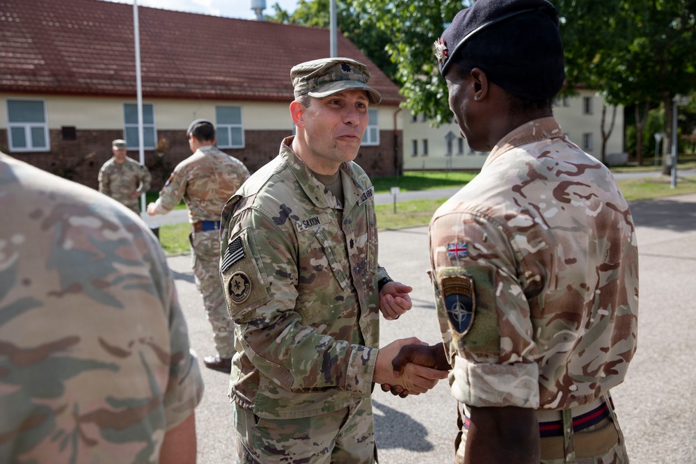 Two military personnel from different nations are shaking hands in what appears to be a formal greeting or ceremony at a military installation. The central figure wears US Army multicam uniform with a lieutenant colonel oak leaf insignia, while the other soldier wears British Army MTP (Multi-Terrain Pattern) uniform with a Union Jack patch and NATO patch. Additional military personnel are visible in the background near institutional buildings.