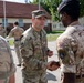 Two military personnel from different nations are shaking hands in what appears to be a formal greeting or ceremony at a military installation. The central figure wears US Army multicam uniform with a lieutenant colonel oak leaf insignia, while the other soldier wears British Army MTP (Multi-Terrain Pattern) uniform with a Union Jack patch and NATO patch. Additional military personnel are visible in the background near institutional buildings.