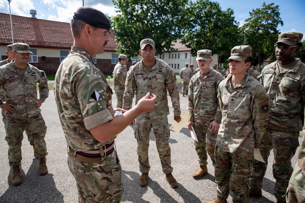 A military briefing or address is taking place outdoors on a paved area, likely a military installation. A senior officer wearing a black beret and British-pattern MTP (Multi-Terrain Pattern) camouflage uniform is speaking to a group of U.S. Army soldiers in OCP (Operational Camouflage Pattern) uniforms. The setting and mixed uniforms suggest a multinational NATO or coalition training exercise or event.