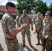 A military briefing or address is taking place outdoors on a paved area, likely a military installation. A senior officer wearing a black beret and British-pattern MTP (Multi-Terrain Pattern) camouflage uniform is speaking to a group of U.S. Army soldiers in OCP (Operational Camouflage Pattern) uniforms. The setting and mixed uniforms suggest a multinational NATO or coalition training exercise or event.