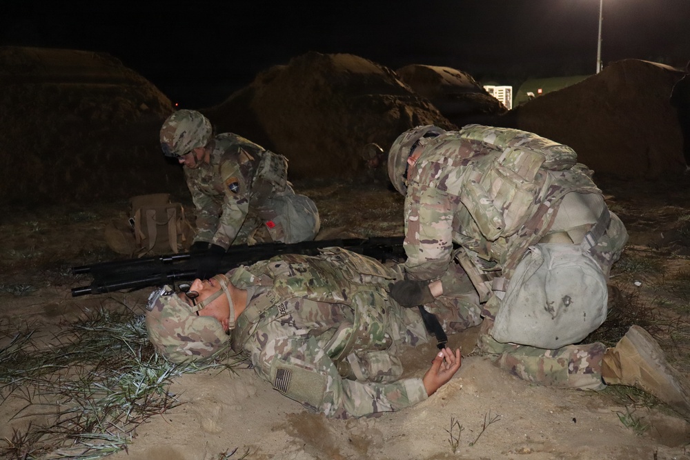 A nighttime military training exercise showing soldiers in Operational Camouflage Pattern (OCP) uniforms conducting tactical combat casualty care (TCCC) or medical training. One soldier is lying on the ground simulating a casualty while another kneels over them performing a simulated medical assessment or treatment. A third soldier is positioned nearby, appearing to be on a radio or observing.