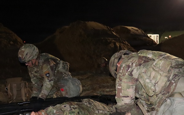 A nighttime military training exercise showing soldiers in Operational Camouflage Pattern (OCP) uniforms conducting tactical combat casualty care (TCCC) or medical training. One soldier is lying on the ground simulating a casualty while another kneels over them performing a simulated medical assessment or treatment. A third soldier is positioned nearby, appearing to be on a radio or observing.