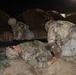 A nighttime military training exercise showing soldiers in Operational Camouflage Pattern (OCP) uniforms conducting tactical combat casualty care (TCCC) or medical training. One soldier is lying on the ground simulating a casualty while another kneels over them performing a simulated medical assessment or treatment. A third soldier is positioned nearby, appearing to be on a radio or observing.