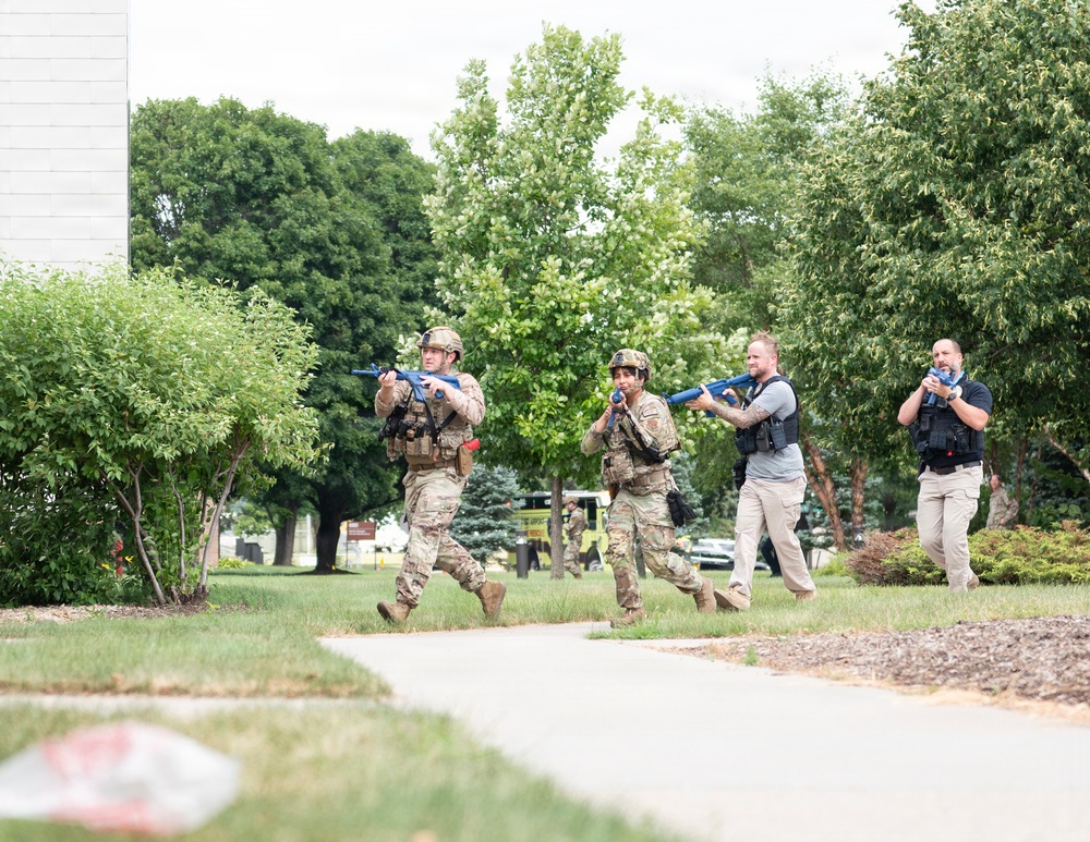 The image shows four individuals advancing in formation along a pathway in a landscaped outdoor area. Two are wearing military-style camouflage uniforms and helmets, while two are in civilian tactical attire. All four are carrying bright blue training/simulation firearms, strongly indicating this is a training exercise or drill rather than a live operation.