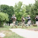 The image shows four individuals advancing in formation along a pathway in a landscaped outdoor area. Two are wearing military-style camouflage uniforms and helmets, while two are in civilian tactical attire. All four are carrying bright blue training/simulation firearms, strongly indicating this is a training exercise or drill rather than a live operation.