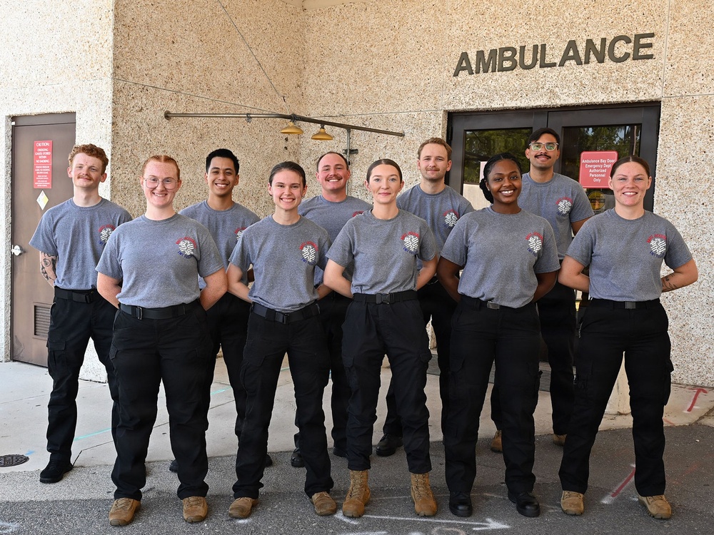 A group of ten individuals in matching uniforms pose for a group photo in front of an ambulance bay entrance. They are wearing identical gray t-shirts with a logo on the chest and black tactical pants, suggesting they are EMS trainees or emergency medical personnel. The setting is the exterior of a medical facility with 'AMBULANCE' signage clearly visible above them.