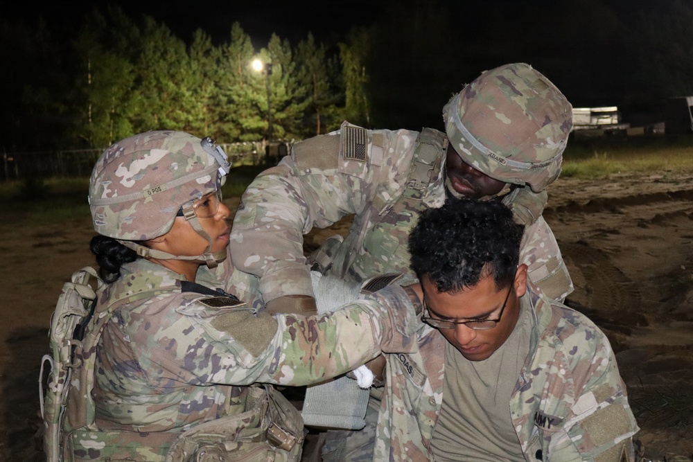 Three U.S. Army soldiers are engaged in what appears to be a nighttime field medical training exercise. Two soldiers in full combat gear and OCP (Operational Camouflage Pattern) uniforms are attending to a third soldier who appears to be acting as a simulated casualty. The scene takes place outdoors at night, illuminated by artificial lighting, with trees visible in the background.