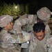 Three U.S. Army soldiers are engaged in what appears to be a nighttime field medical training exercise. Two soldiers in full combat gear and OCP (Operational Camouflage Pattern) uniforms are attending to a third soldier who appears to be acting as a simulated casualty. The scene takes place outdoors at night, illuminated by artificial lighting, with trees visible in the background.
