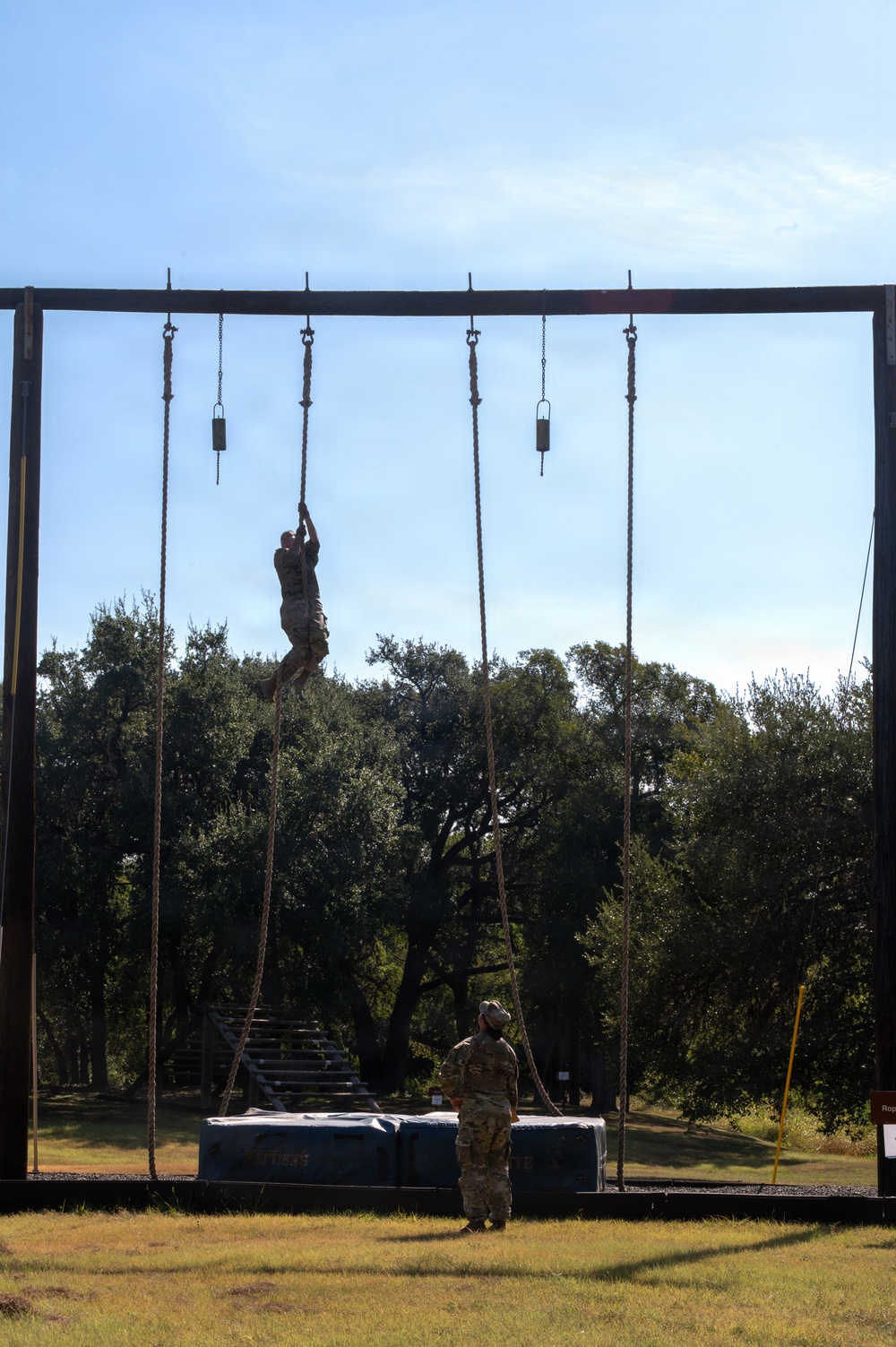 A military obstacle course rope climb exercise is underway, with one soldier actively climbing a rope near the top of a tall metal frame structure while a second soldier stands below on a platform observing or spotting. The setting appears to be an outdoor military training facility surrounded by trees under a clear blue sky. Multiple ropes with weighted ends hang from the overhead crossbar, indicating this is a dedicated rope climb training station.