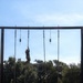 A military obstacle course rope climb exercise is underway, with one soldier actively climbing a rope near the top of a tall metal frame structure while a second soldier stands below on a platform observing or spotting. The setting appears to be an outdoor military training facility surrounded by trees under a clear blue sky. Multiple ropes with weighted ends hang from the overhead crossbar, indicating this is a dedicated rope climb training station.