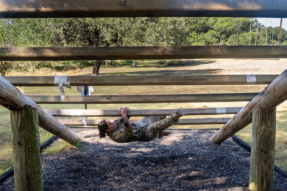 A soldier in camouflage uniform is navigating a military obstacle course, performing an inverted crawl or monkey-bar-style traverse underneath a horizontal wooden ladder structure. The individual is lying on their back and pulling themselves along the wooden rails using their hands and legs. The setting appears to be an outdoor military training facility with gravel ground cover and wooded surroundings.