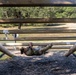 A soldier in camouflage uniform is navigating a military obstacle course, performing an inverted crawl or monkey-bar-style traverse underneath a horizontal wooden ladder structure. The individual is lying on their back and pulling themselves along the wooden rails using their hands and legs. The setting appears to be an outdoor military training facility with gravel ground cover and wooded surroundings.