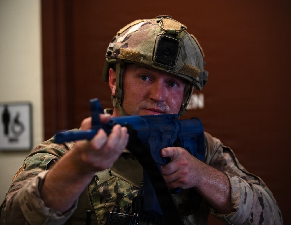 A military or law enforcement personnel in full tactical gear is holding a blue training/simulation firearm in a ready position. The individual is wearing MultiCam/OCP camouflage uniform, a tactical helmet with a mounted camera device, and body armor. The setting appears to be an indoor facility, likely during a training exercise, as indicated by the blue-colored inert training weapon.