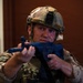 A military or law enforcement personnel in full tactical gear is holding a blue training/simulation firearm in a ready position. The individual is wearing MultiCam/OCP camouflage uniform, a tactical helmet with a mounted camera device, and body armor. The setting appears to be an indoor facility, likely during a training exercise, as indicated by the blue-colored inert training weapon.