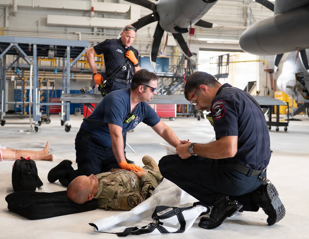A medical training exercise or emergency response drill is taking place inside an aircraft hangar. Two first responders in dark blue uniforms are attending to a person in military camouflage clothing lying on the hangar floor, while a third responder approaches from the background. The setting includes a large military or civilian aircraft with visible propeller blades overhead.
