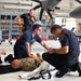 A medical training exercise or emergency response drill is taking place inside an aircraft hangar. Two first responders in dark blue uniforms are attending to a person in military camouflage clothing lying on the hangar floor, while a third responder approaches from the background. The setting includes a large military or civilian aircraft with visible propeller blades overhead.