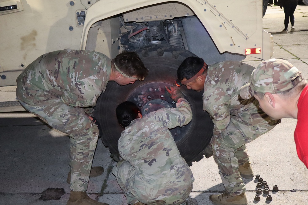 Four individuals in U.S. Army OCP (Operational Camouflage Pattern) uniforms are performing a tire change or wheel installation on a military tactical vehicle, likely an MRAP or HMMWV. Three soldiers are actively handling a large military-grade tire against the wheel hub, while a fourth observes from the right. Lug nuts are visibly staged on the ground to the right, indicating the task is in progress.