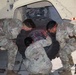 Four individuals in U.S. Army OCP (Operational Camouflage Pattern) uniforms are performing a tire change or wheel installation on a military tactical vehicle, likely an MRAP or HMMWV. Three soldiers are actively handling a large military-grade tire against the wheel hub, while a fourth observes from the right. Lug nuts are visibly staged on the ground to the right, indicating the task is in progress.