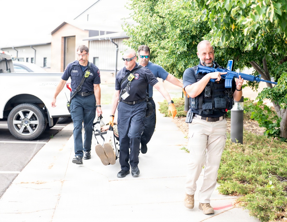 The image shows what appears to be a training exercise involving law enforcement and emergency medical personnel. Four individuals in uniform are moving along a sidewalk, with one armed person in tactical gear carrying a blue-colored rifle (indicating a training/simulation weapon) while two others carry what appears to be a stretcher or litter with a simulated casualty. The blue color of the firearm and the coordinated movement strongly suggest this is a mass casualty or active shooter response drill.