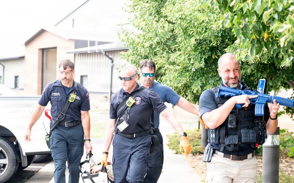 The image shows what appears to be a training exercise involving law enforcement and emergency medical personnel. Four individuals in uniform are moving along a sidewalk, with one armed person in tactical gear carrying a blue-colored rifle (indicating a training/simulation weapon) while two others carry what appears to be a stretcher or litter with a simulated casualty. The blue color of the firearm and the coordinated movement strongly suggest this is a mass casualty or active shooter response drill.