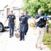 The image shows what appears to be a training exercise involving law enforcement and emergency medical personnel. Four individuals in uniform are moving along a sidewalk, with one armed person in tactical gear carrying a blue-colored rifle (indicating a training/simulation weapon) while two others carry what appears to be a stretcher or litter with a simulated casualty. The blue color of the firearm and the coordinated movement strongly suggest this is a mass casualty or active shooter response drill.