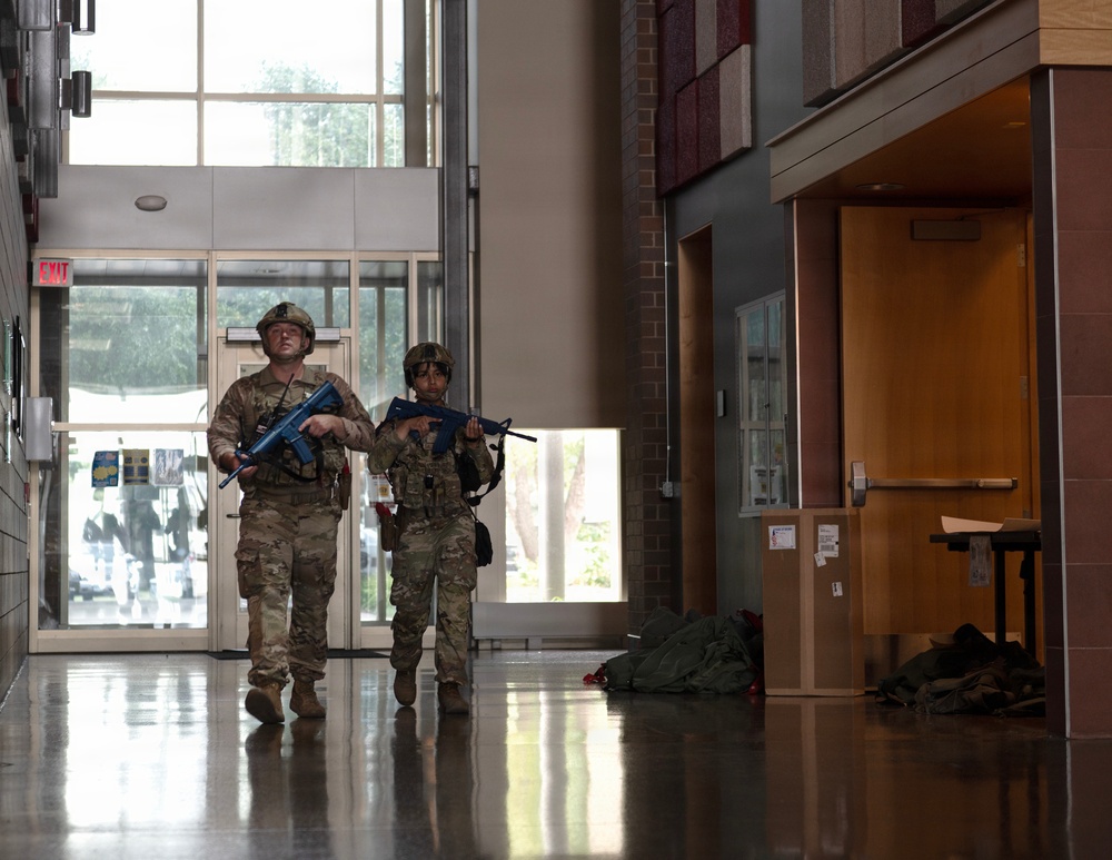 Two individuals in military/tactical camouflage uniforms and helmets are moving through what appears to be a building lobby or corridor, carrying blue-colored training firearms. The blue color of the weapons indicates these are simunition or training weapons, strongly suggesting this is a training exercise or drill rather than an actual operation. The setting appears to be a civilian building such as a school or government facility.