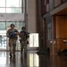 Two individuals in military/tactical camouflage uniforms and helmets are moving through what appears to be a building lobby or corridor, carrying blue-colored training firearms. The blue color of the weapons indicates these are simunition or training weapons, strongly suggesting this is a training exercise or drill rather than an actual operation. The setting appears to be a civilian building such as a school or government facility.