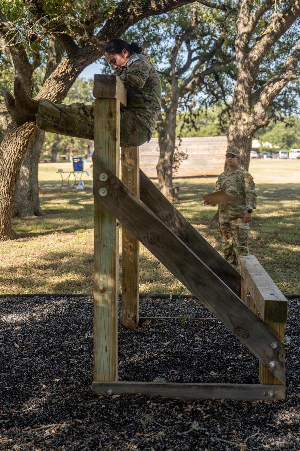 A military service member in OCP (Operational Camouflage Pattern) uniform is navigating a wooden obstacle course structure, appearing to climb over or mount a vertical post with an angled ramp. A second uniformed service member stands in the background holding a clipboard, likely serving as an evaluator or grader. The setting appears to be an outdoor military training area with large oak trees and rubber mulch ground cover.