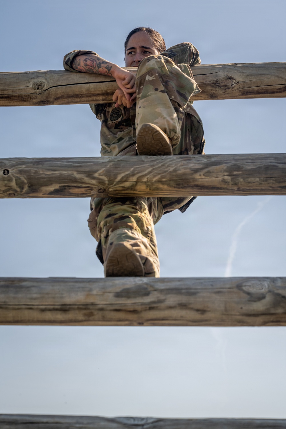 A female soldier in MultiCam/OCP camouflage uniform is climbing over a wooden log obstacle, likely part of a military obstacle course or physical training exercise. She is captured mid-climb, with her arms hooked over the top log rail and her legs navigating between the lower rails. The low-angle shot looking upward emphasizes the height and physical effort of the task.