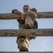 A female soldier in MultiCam/OCP camouflage uniform is climbing over a wooden log obstacle, likely part of a military obstacle course or physical training exercise. She is captured mid-climb, with her arms hooked over the top log rail and her legs navigating between the lower rails. The low-angle shot looking upward emphasizes the height and physical effort of the task.