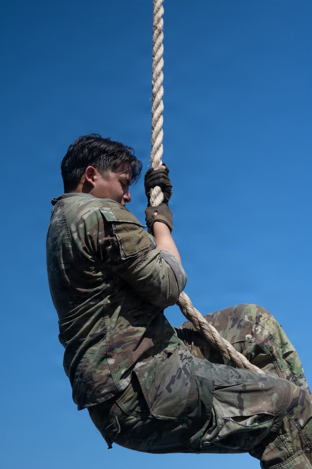 A military personnel in MultiCam/OCP camouflage uniform is climbing a thick rope against a clear blue sky. The individual is wearing tactical gloves and using both hands and legs to grip the rope in a classic rope-climb technique. The low-angle shot emphasizes the height and physical effort of the exercise.