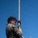 A military personnel in MultiCam/OCP camouflage uniform is climbing a thick rope against a clear blue sky. The individual is wearing tactical gloves and using both hands and legs to grip the rope in a classic rope-climb technique. The low-angle shot emphasizes the height and physical effort of the exercise.