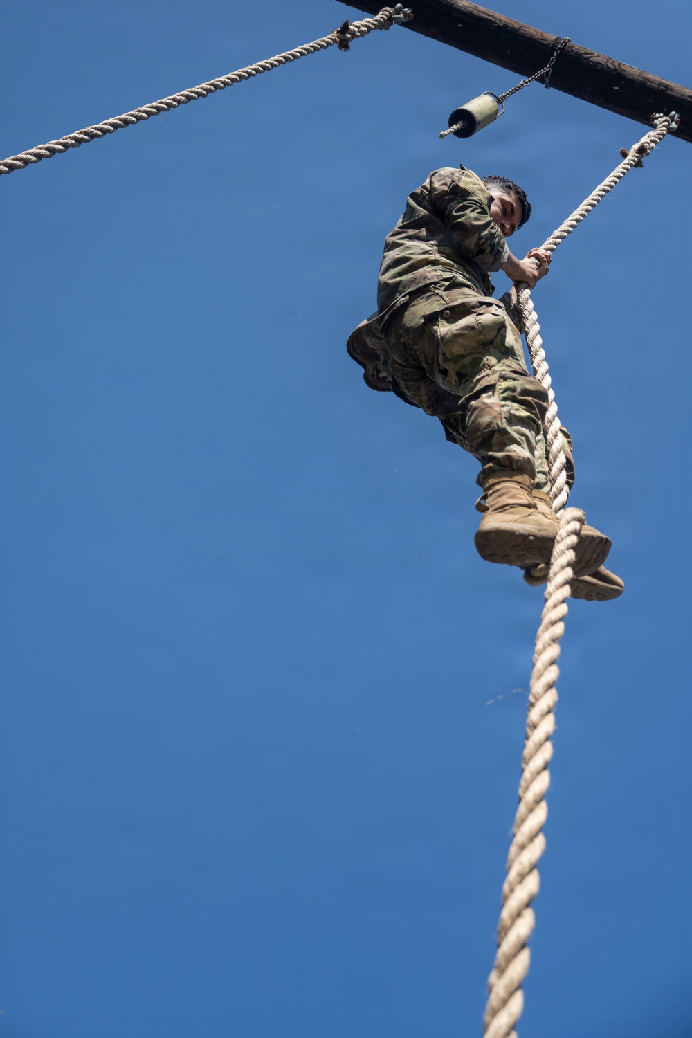 A soldier in military camouflage uniform and tan combat boots is climbing a thick rope on an outdoor obstacle course. The photo is taken from a low angle looking upward, with a clear blue sky as the background. The individual has reached near the top of the rope, close to a wooden overhead structure.