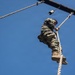A soldier in military camouflage uniform and tan combat boots is climbing a thick rope on an outdoor obstacle course. The photo is taken from a low angle looking upward, with a clear blue sky as the background. The individual has reached near the top of the rope, close to a wooden overhead structure.
