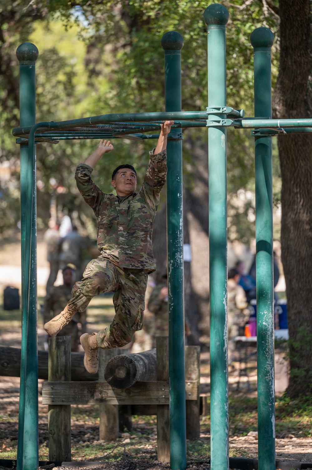 A soldier in camouflage uniform is navigating a military obstacle course, specifically traversing horizontal monkey bars or overhead ladder rungs. The setting appears to be an outdoor military training facility with trees providing shade. Other personnel in military attire are visible in the background observing or waiting.