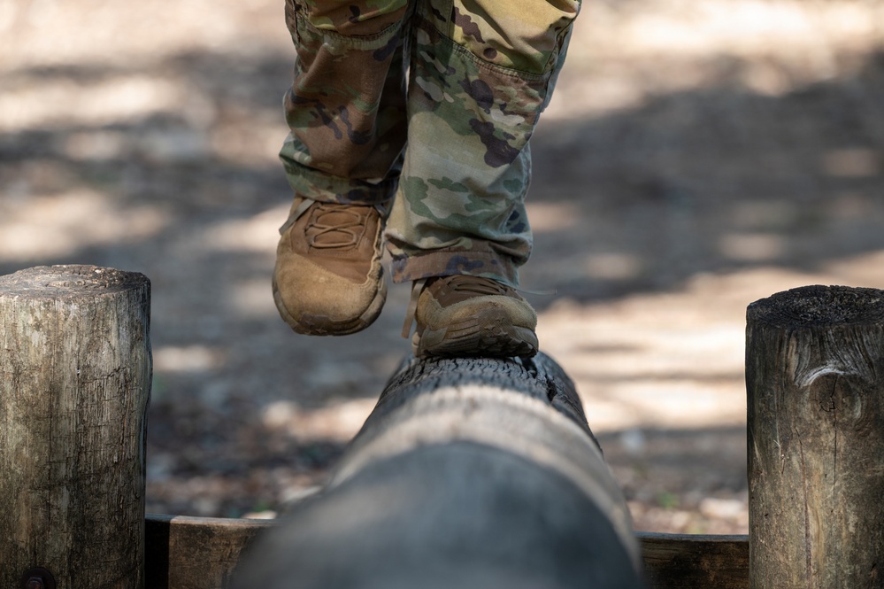 A person wearing military camouflage uniform and tan combat boots is navigating a balance beam obstacle, consisting of a horizontal log supported by vertical wooden posts. The image is captured from a low angle focusing on the lower legs and feet. This appears to be a military or fitness obstacle course set in an outdoor wooded environment.