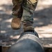 A person wearing military camouflage uniform and tan combat boots is navigating a balance beam obstacle, consisting of a horizontal log supported by vertical wooden posts. The image is captured from a low angle focusing on the lower legs and feet. This appears to be a military or fitness obstacle course set in an outdoor wooded environment.