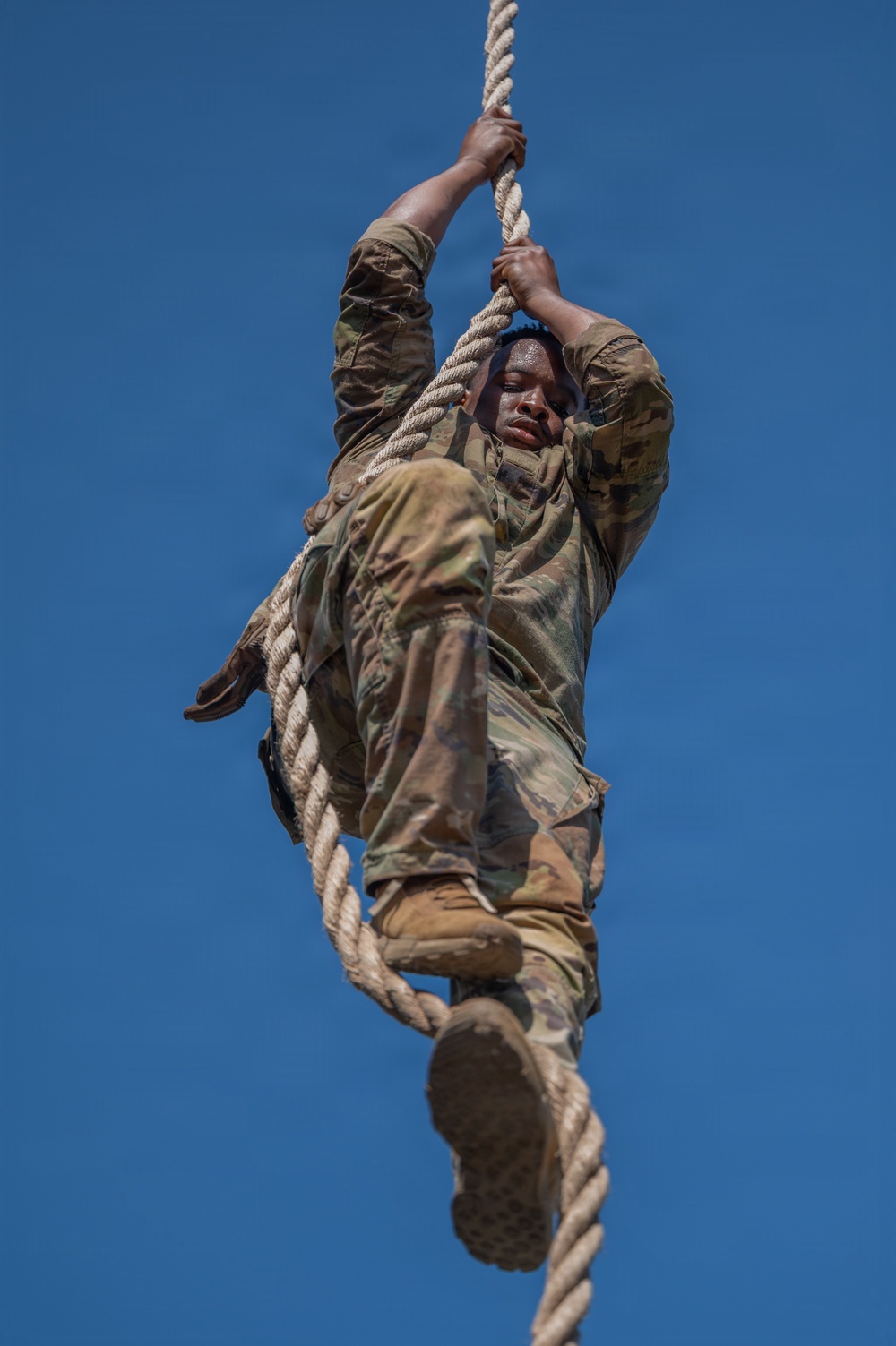 A soldier in OCP (Operational Camouflage Pattern) uniform is climbing a thick rope against a clear blue sky background. The image is taken from a low angle looking upward, capturing the soldier mid-climb with both hands gripping the rope and feet wrapped around it. The photograph appears to document a military physical training or obstacle course exercise.