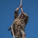 A soldier in OCP (Operational Camouflage Pattern) uniform is climbing a thick rope against a clear blue sky background. The image is taken from a low angle looking upward, capturing the soldier mid-climb with both hands gripping the rope and feet wrapped around it. The photograph appears to document a military physical training or obstacle course exercise.