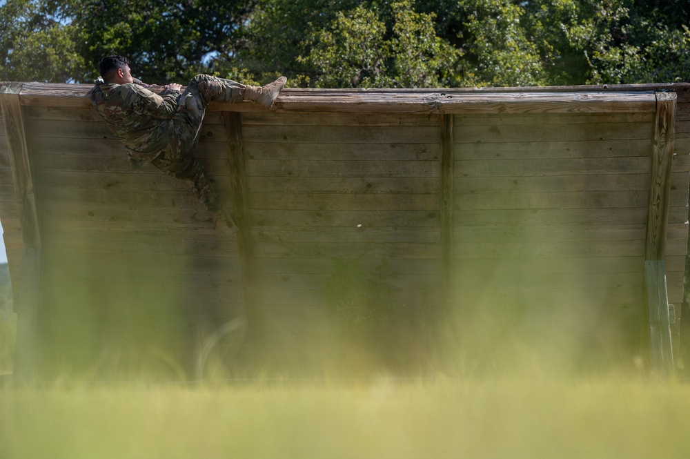 A soldier in military camouflage uniform is scaling or climbing over a tall wooden wall obstacle, likely part of a military training course or obstacle course. The person is positioned at the top of the wall with their body draped over it, wearing combat boots and full military attire. The foreground features blurred green grass, suggesting the photo was taken from a low angle through vegetation.