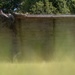 A soldier in military camouflage uniform is scaling or climbing over a tall wooden wall obstacle, likely part of a military training course or obstacle course. The person is positioned at the top of the wall with their body draped over it, wearing combat boots and full military attire. The foreground features blurred green grass, suggesting the photo was taken from a low angle through vegetation.