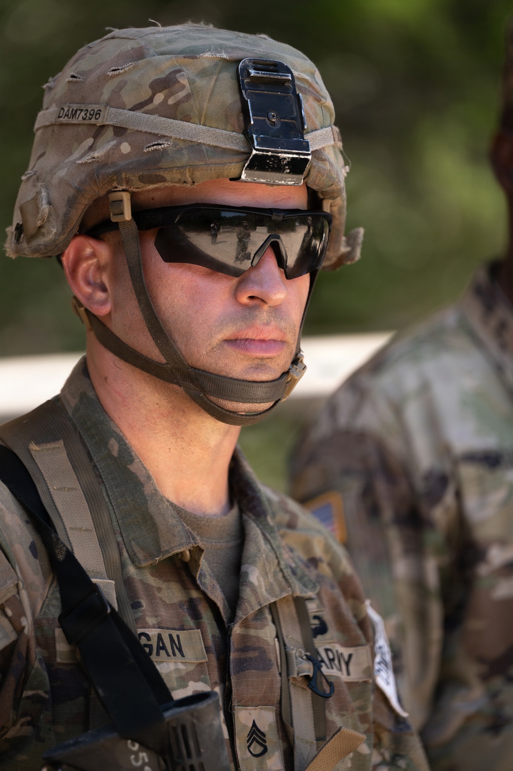 A U.S. Army soldier is photographed in close-up wearing Operational Camouflage Pattern (OCP) uniform and helmet with tactical eyewear. The soldier's name tape reads 'GAN' (partially visible) and rank insignia indicates Staff Sergeant (SSG). The helmet bears the identifier 'DAM7396' and has a night-vision mount bracket attached to the front.