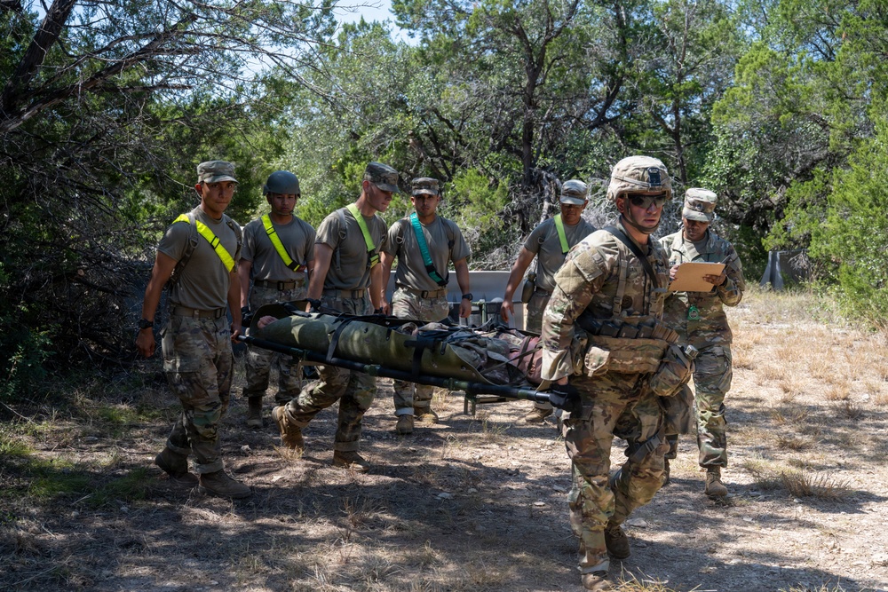 A group of military personnel in uniform are conducting a casualty evacuation (CASEVAC) training exercise in an outdoor wooded area. Several soldiers are carrying a simulated casualty on a litter/stretcher along a dirt path, while a soldier in full combat gear leads the group and another appears to be evaluating or documenting the exercise on a clipboard. The setting appears to be a training environment based on the organized, supervised nature of the activity.
