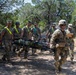 A group of military personnel in uniform are conducting a casualty evacuation (CASEVAC) training exercise in an outdoor wooded area. Several soldiers are carrying a simulated casualty on a litter/stretcher along a dirt path, while a soldier in full combat gear leads the group and another appears to be evaluating or documenting the exercise on a clipboard. The setting appears to be a training environment based on the organized, supervised nature of the activity.
