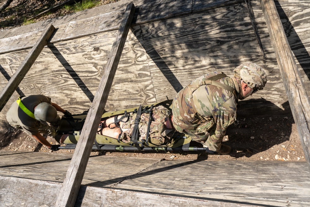 Two military personnel in camouflage uniforms are maneuvering a litter (military stretcher) with a simulated casualty through a confined wooden structure, likely during a combat casualty care training exercise. The photo is taken from an elevated overhead angle, showing the soldiers navigating the stretcher through a narrow opening between wooden planks. This appears to be a tactical combat casualty care (TCCC) or field medical training scenario.