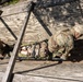 Two military personnel in camouflage uniforms are maneuvering a litter (military stretcher) with a simulated casualty through a confined wooden structure, likely during a combat casualty care training exercise. The photo is taken from an elevated overhead angle, showing the soldiers navigating the stretcher through a narrow opening between wooden planks. This appears to be a tactical combat casualty care (TCCC) or field medical training scenario.