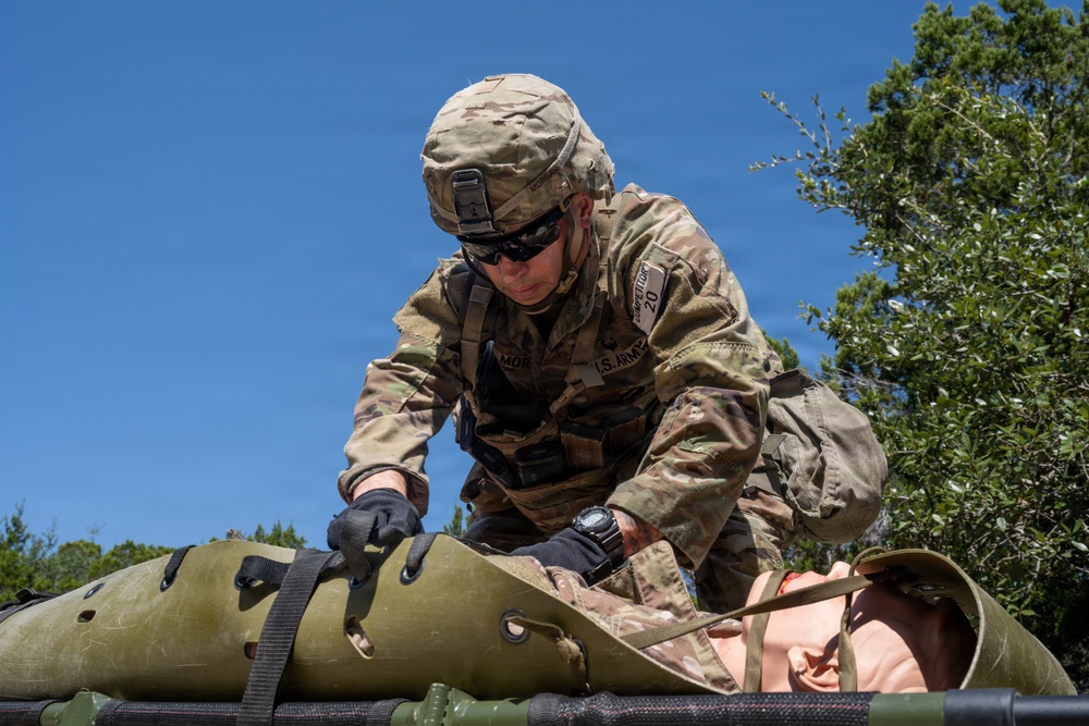 A U.S. Army soldier in full OCP (Operational Camouflage Pattern) uniform is working with a litter/stretcher, appearing to secure or adjust straps on what may be a casualty or training mannequin. The soldier is wearing a combat helmet, tactical sunglasses, and black gloves, with a name tape partially visible reading 'MOR' and a competition number patch '20' on the sleeve. The setting appears to be an outdoor training exercise under clear blue skies.