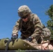 A U.S. Army soldier in full OCP (Operational Camouflage Pattern) uniform is working with a litter/stretcher, appearing to secure or adjust straps on what may be a casualty or training mannequin. The soldier is wearing a combat helmet, tactical sunglasses, and black gloves, with a name tape partially visible reading 'MOR' and a competition number patch '20' on the sleeve. The setting appears to be an outdoor training exercise under clear blue skies.